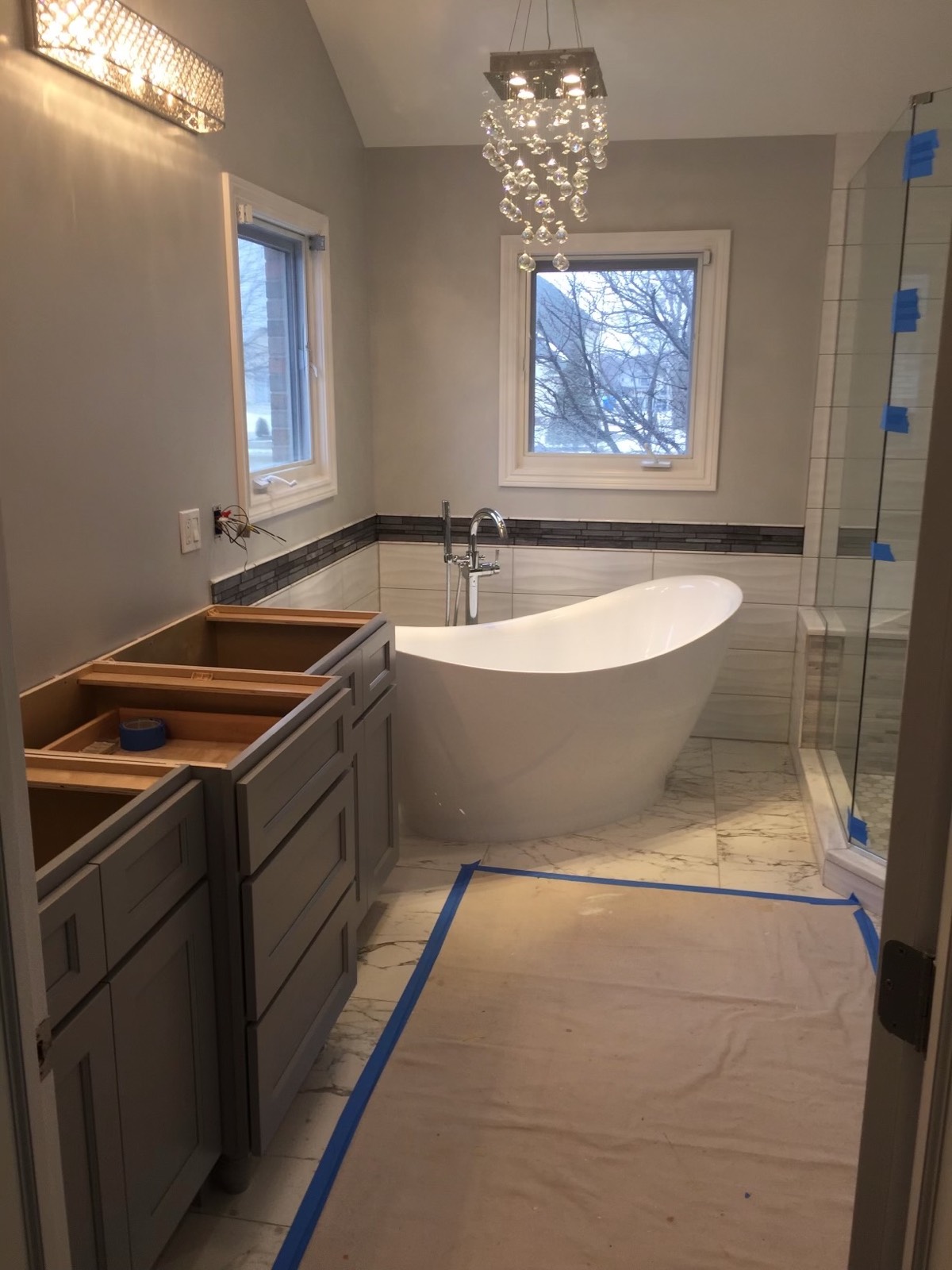 Primary bath mid-install: gray vanity cabinets set before the tops, a freestanding slipper tub and crystal chandelier, floor protected