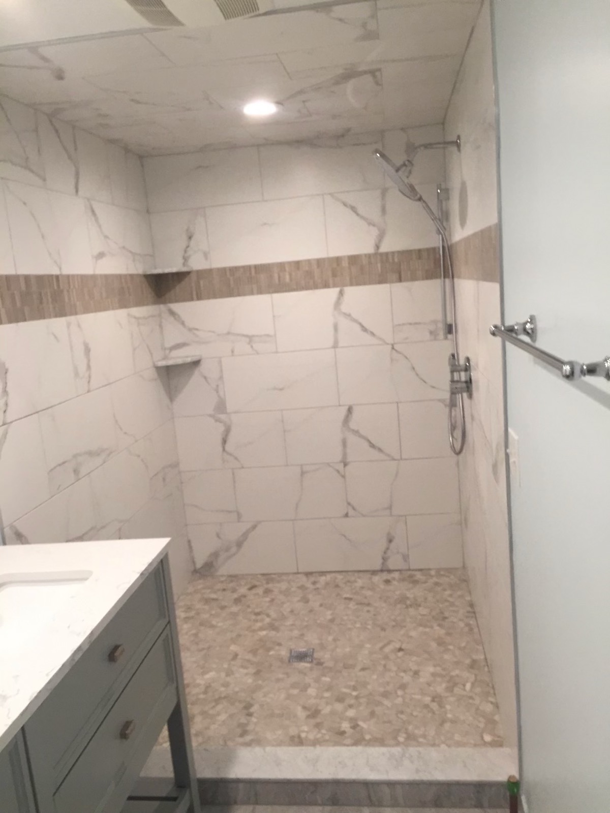 Large walk-in shower in marble-look porcelain with a mosaic band, corner shelves and a pebble floor