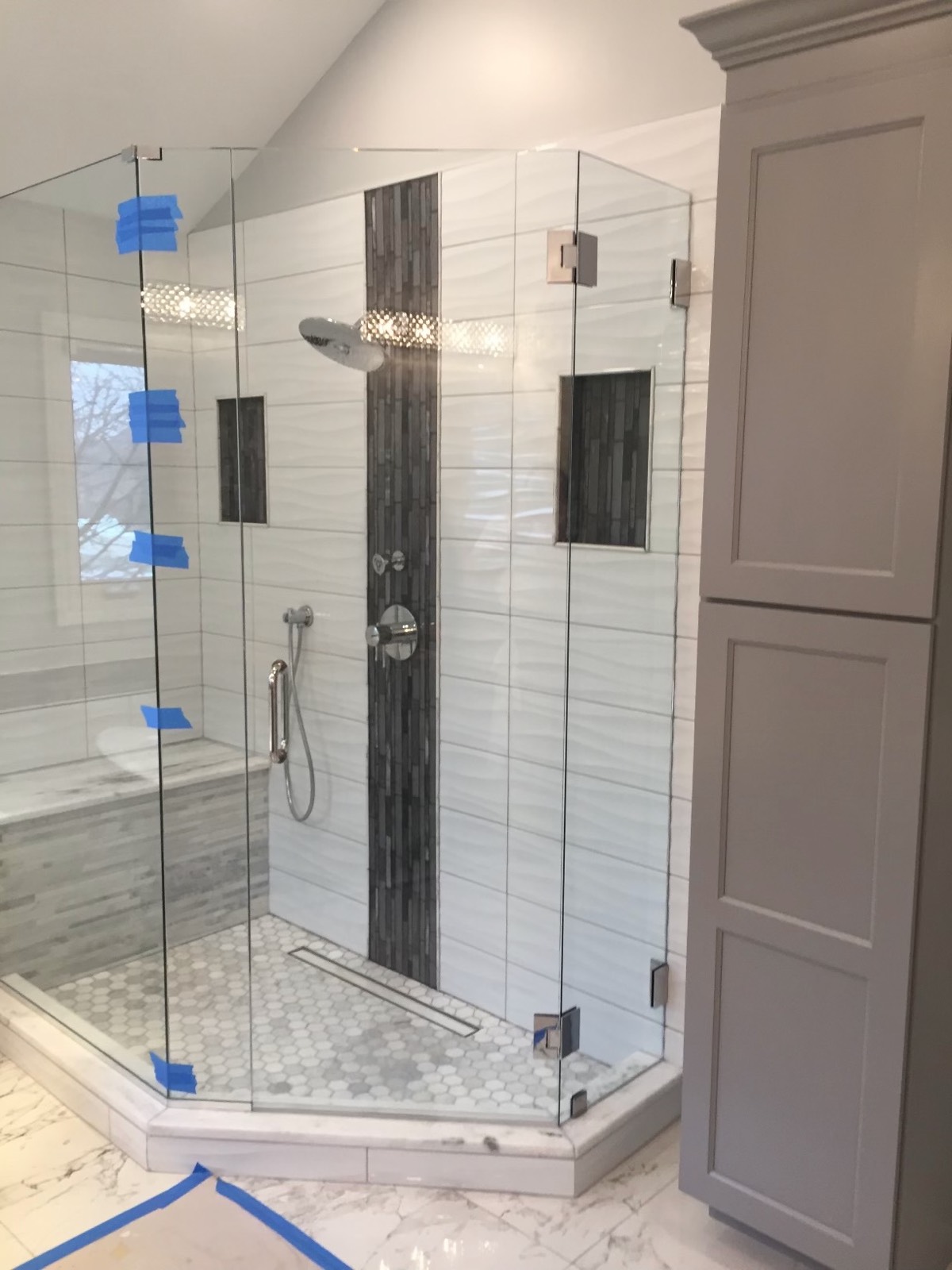 Neo-angle frameless glass shower with a vertical mosaic stripe, bench, linear drain and a gray linen tower